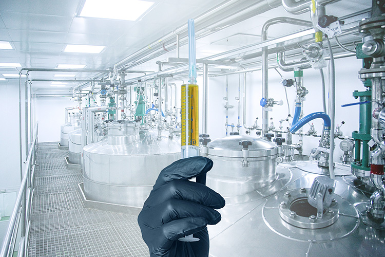 Gloved hand holding syringe in front of biomanufacturing tanks
